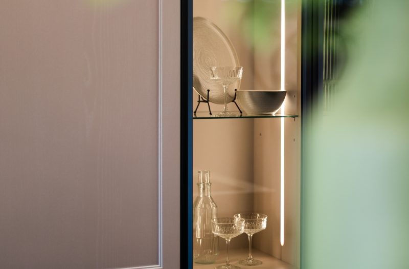 Detail of a Nolte Lugano Lack kitchen display in Amersham featuring an illuminated glass cabinet styled with glassware and tableware.