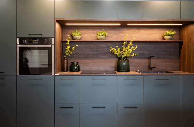 Nolte Manhattan Uni kitchen display at the Amersham showroom with green cabinetry, warm wood accents, integrated oven and recessed task lighting.