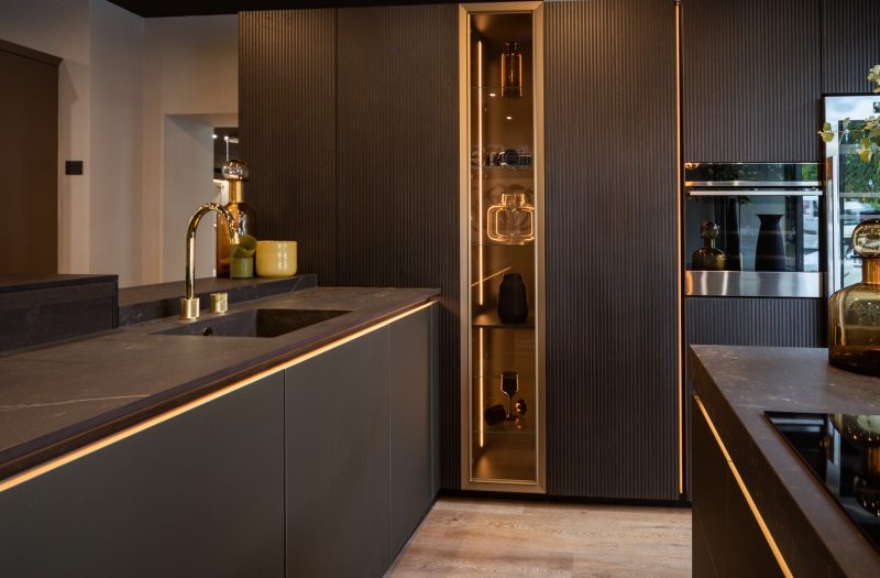Nolte neoLodge kitchen display in Amersham featuring dark fluted cabinetry, integrated appliances, illuminated glass storage and warm accent lighting.