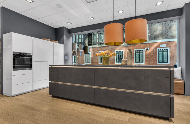 Nolte Kitchens Westminster showroom featuring a contemporary dark kitchen island with white handleless cabinetry, integrated appliances and large windows overlooking London.