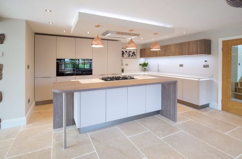 Contemporary Nolte Feel and Manhattan kitchen featuring soft neutral cabinetry, wood-effect accents, a large island with integrated hob, pendant lighting and natural stone flooring.