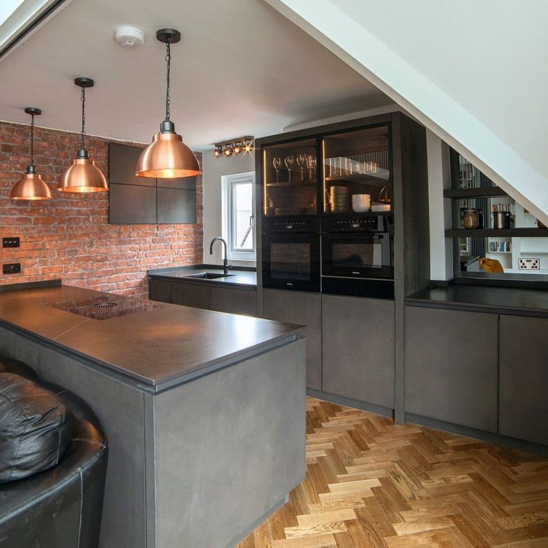 Contemporary Nolte Portland Cement Anthracite kitchen within a Georgian townhouse, featuring dark cabinetry, an island, exposed brickwork, copper pendant lights and parquet flooring.