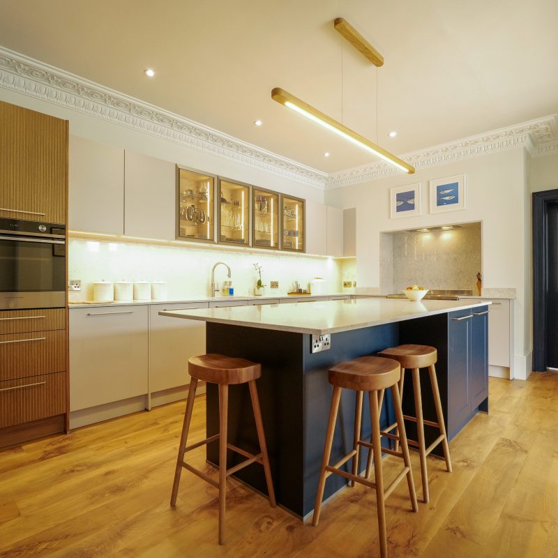 Modern German kitchen with wood-effect cabinetry, light matte cabinets, navy blue island, quartz worktops and warm LED feature lighting.
