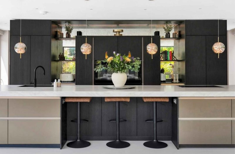 Luxury dark kitchen designed by Nolte Kitchens Henley-on-Thames, featuring neoLODGE fluted cabinetry and neoPURE Bronze finishes, a large island with a white Laminam Taj Mahal worktop, integrated NEFF ovens, illuminated shelving and three breakfast bar stools beneath statement pendant lighting.