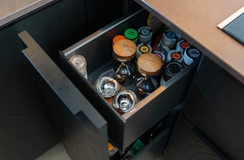 Clever kitchen storage in Rachel's dark neoSLATE kitchen in Henley-on-Thames, featuring an open pull-out internal spice drawer with neatly organised herbs, spices and seasoning jars inside textured black cabinetry.