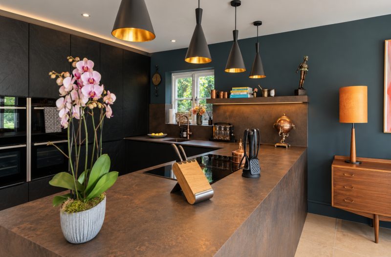 Close-up of Rachel's dark kitchen peninsula in Henley-on-Thames, featuring Laminam Ossido Bruno porcelain worktops and cladding, a flush-fitting induction hob, textured black neoSLATE cabinetry and warm pendant lighting.