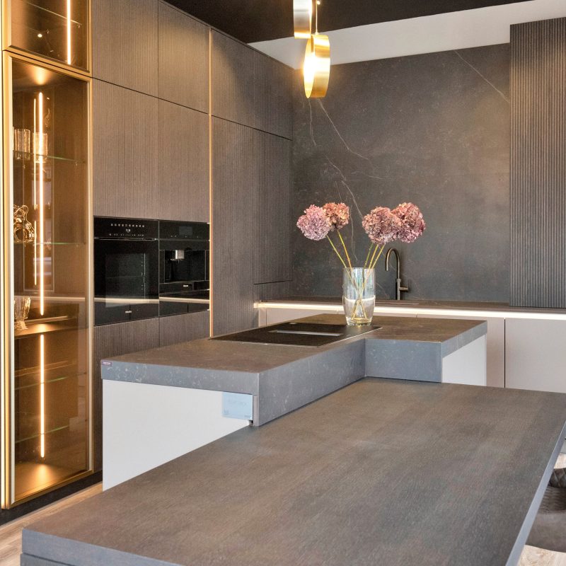 Nolte Kitchens neoLodge in Blackened Oak kitchen display at the Henley-on-Thames showroom, featuring floor-to-ceiling ribbed cabinetry with brushed gold-framed illuminated glass display cabinets, a dark stone-effect worktop and matching dining table, induction hob with integrated sink, dark marble-effect splashback, integrated oven and coffee machine tower, gold pendant lighting, and a leather dining chair styled with a pink hydrangea floral arrangement.