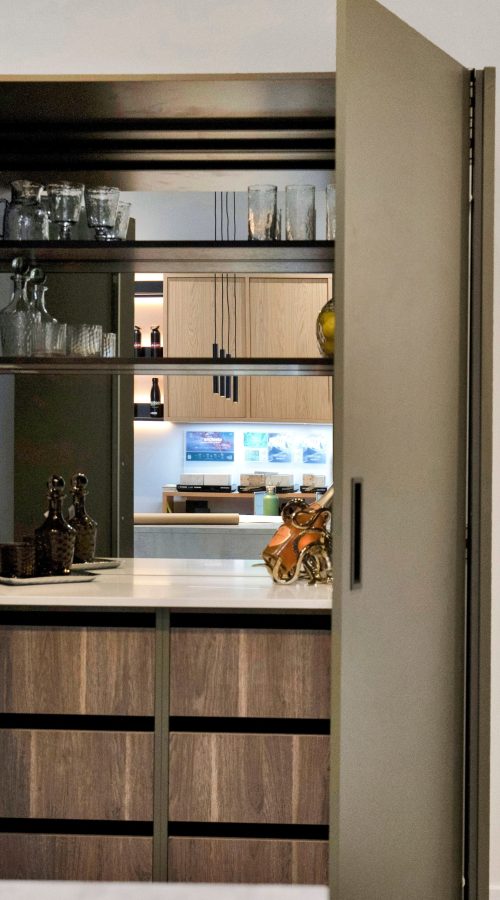 Nolte Kitchens hidden bar display in Soft Lack Olive at the Henley-on-Thames showroom, shown with bi-fold doors open to reveal internal open shelving styled with glassware, a stone-effect worktop, and oak-effect drawer cabinetry, with a Laminam superior natural surfaces sample stand displaying porcelain worktop finishes in the foreground.