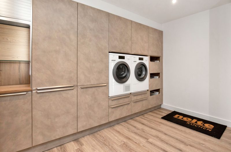 Nolte Kitchens hidden utility room display in Portland Cement Agate Grey at the Henley-on-Thames showroom, featuring floor-to-ceiling handleless cabinetry with bar pulls, integrated side-by-side Miele washing machine and tumble dryer with open storage niches, a tambour roller shutter cabinet with oak-effect interior and timber worktop, and light oak flooring with a Nolte Küchen branded doormat.
