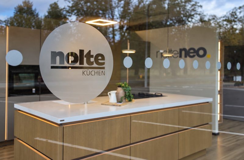View through the Nolte Wakefield showroom window of a neoLine oak kitchen island with a white worktop and frosted Nolte logo