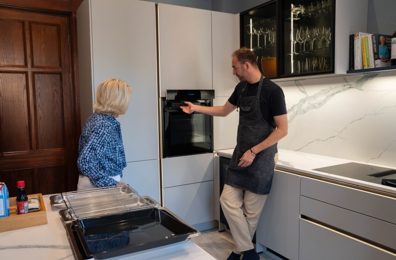 neo Chef Experience demonstrating the built-in oven to Caroline in a Platinum Grey Nolte kitchen with illuminated glass wall cabinets
